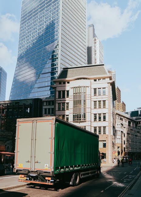 A large green moving truck with a closed cargo area is parked on a city street in front of modern glass skyscrapers and older stone buildings. The truck appears to be preparing for a home relocation or furniture transport as part of a professional moving service. The truck is positioned on the road, with some pedestrians visible nearby and a cityscape backdrop featuring tall office buildings and blue sky. The scene depicts an urban environment during daytime, with the truck likely associated with house removals conducted by Removal Company Mayfair, situated close to high-end commercial and residential areas, supporting the logistics of packing, loading, and transporting furniture and boxes.