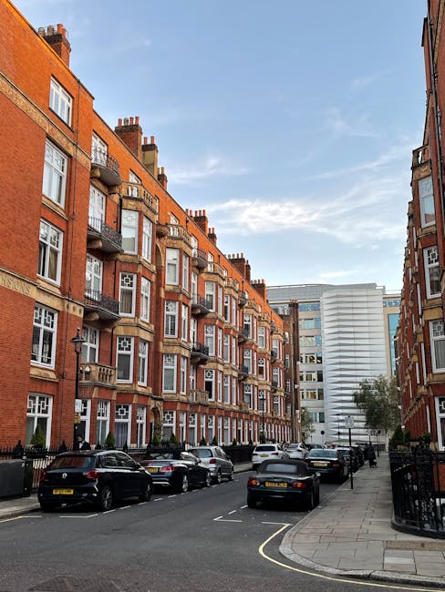 A street scene in a residential area featuring a row of multi-storey red-brick buildings with white-framed bay windows and small balconies, with several cars parked along the curb on a paved road. The sidewalk runs alongside the buildings, with a black wrought iron fence visible on the right and a few small trees planted along the pavement. The sky is partly cloudy with patches of blue. This setting showcases an urban environment typical of high-end housing in Mayfair, where house removals and furniture transport services by Removal Company Mayfair may involve loading large packing boxes, furniture, and appliances into vans parked nearby, with potential interior and exterior packing and moving logistics visible for home relocation projects.