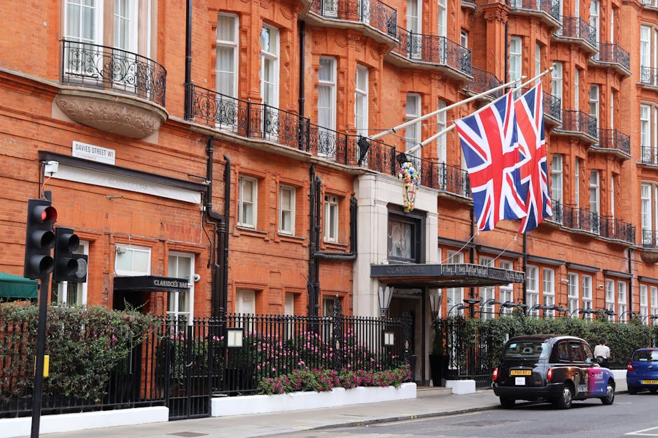 The exterior of The Dorchester hotel in Mayfair, London, featuring a beige stone facade with black and gold accented entrance doors. In front of the hotel, a group of three individuals dressed in form